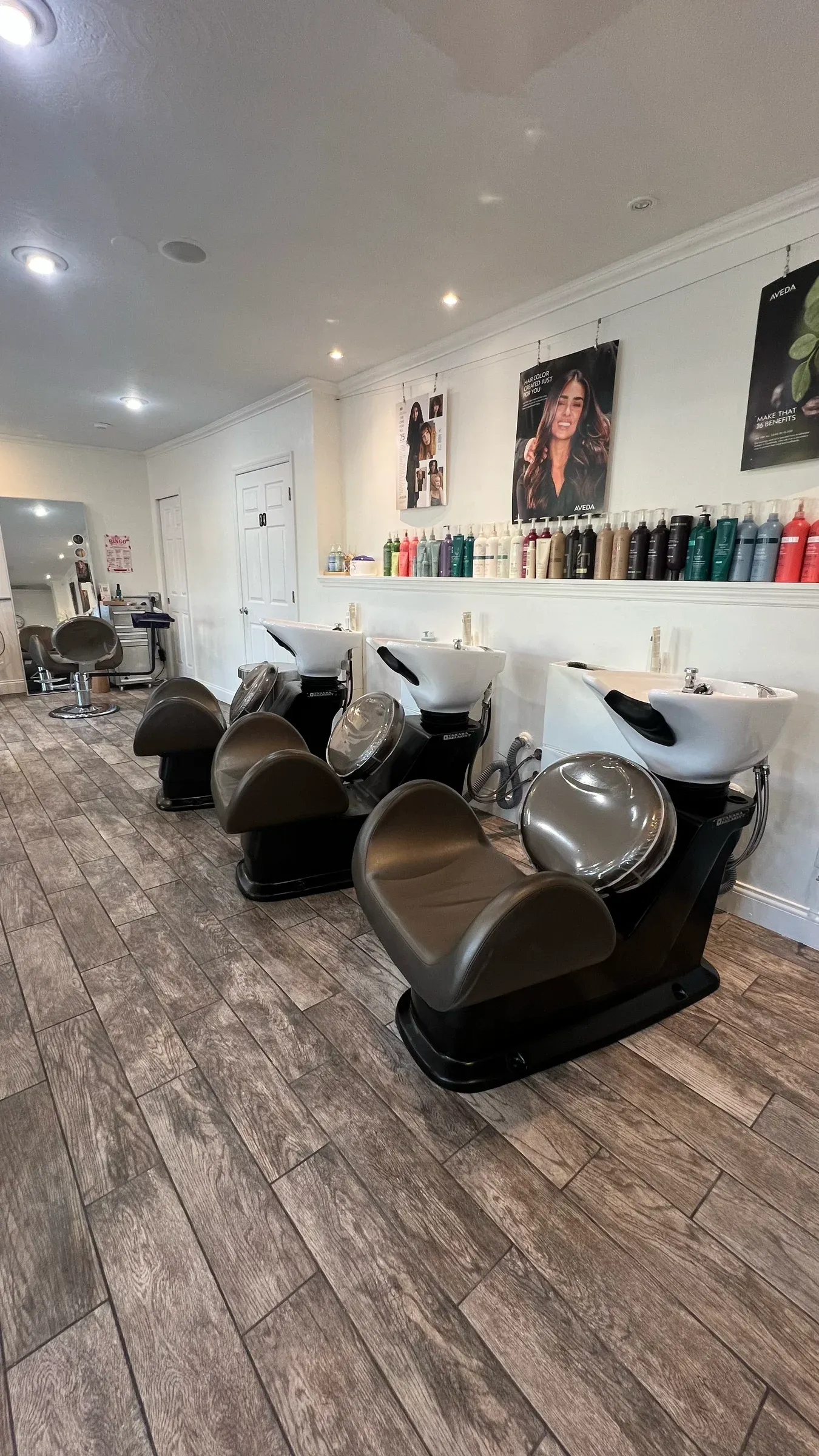 A row of three shampoo bowls with reclining chairs beneath a shelf of Aveda products